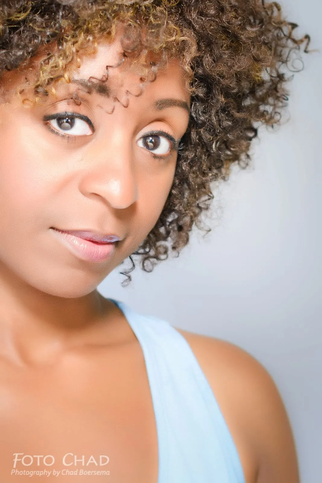 Studio headshot of a woman with curly, honey-highlighted hair in a light-blue top against a grey backdrop, credited Foto Chad, Photography by Chad Boersema.