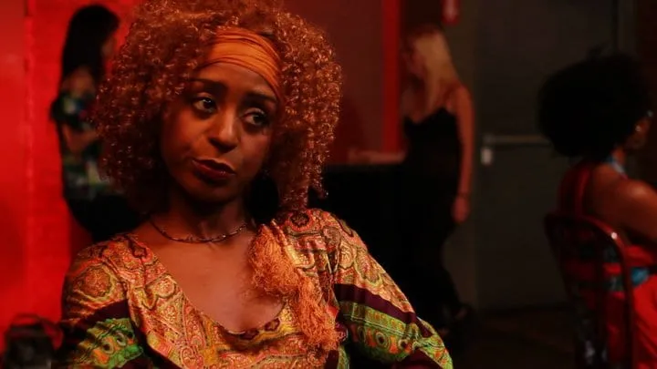 Production still from 12-Steps to Recovery: a woman with curly copper hair and an orange headband glances aside in a red-lit bar.