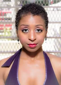 Headshot of a woman with short natural hair, red lipstick and a purple halter top in front of a chain-link fence.