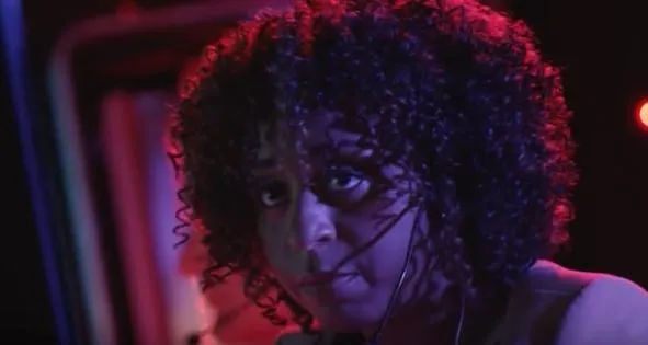 Trailer still from The Fast and the Fierce showing a woman with dark curly hair lit in red and purple light.