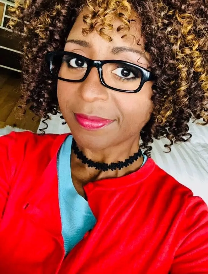 Close-up selfie of a smiling woman with curly brown hair, black-framed glasses, a red top and a black choker.