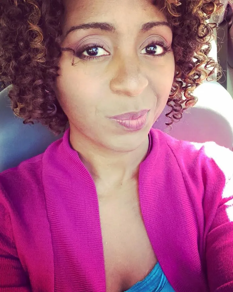 Selfie of a woman with curly brown hair in a magenta cardigan over a teal top, seated in a car.