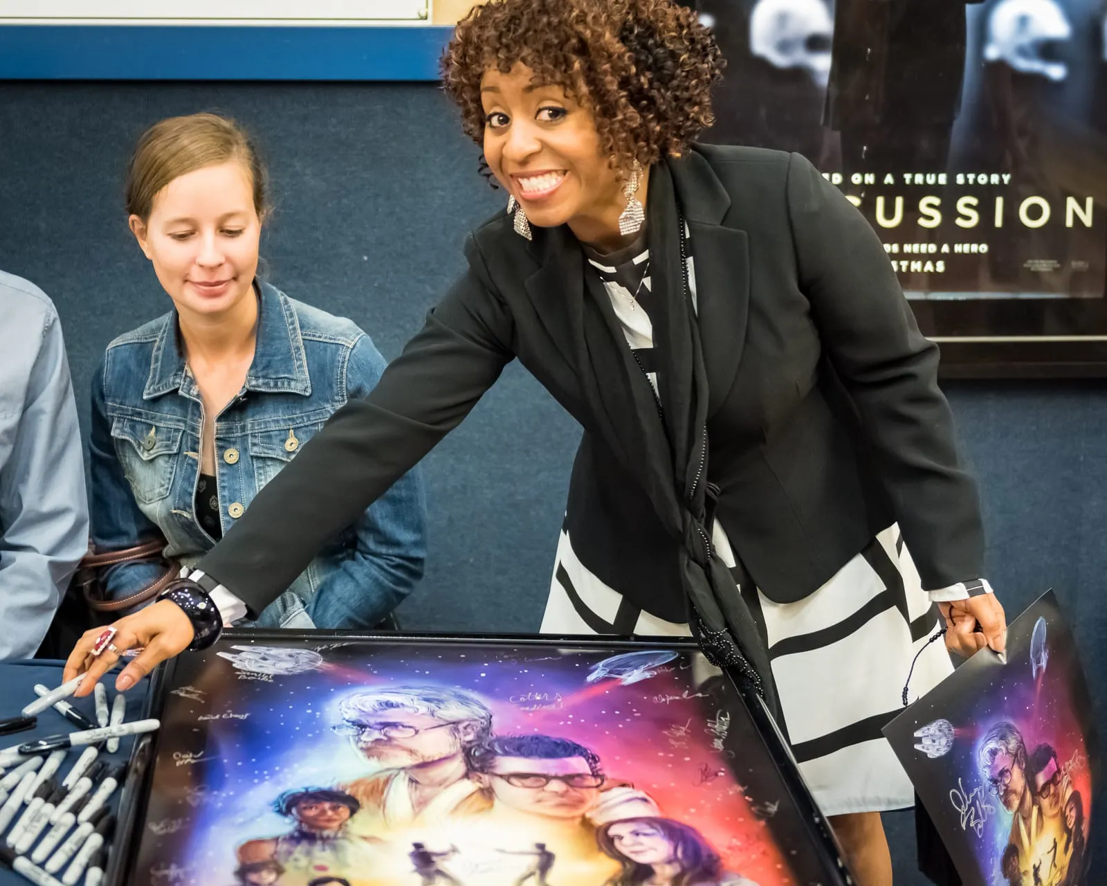 Sophia Thomas smiling as she signs a framed Star Trek Wars poster at the premiere, with markers on the table and a woman in a denim jacket beside her
