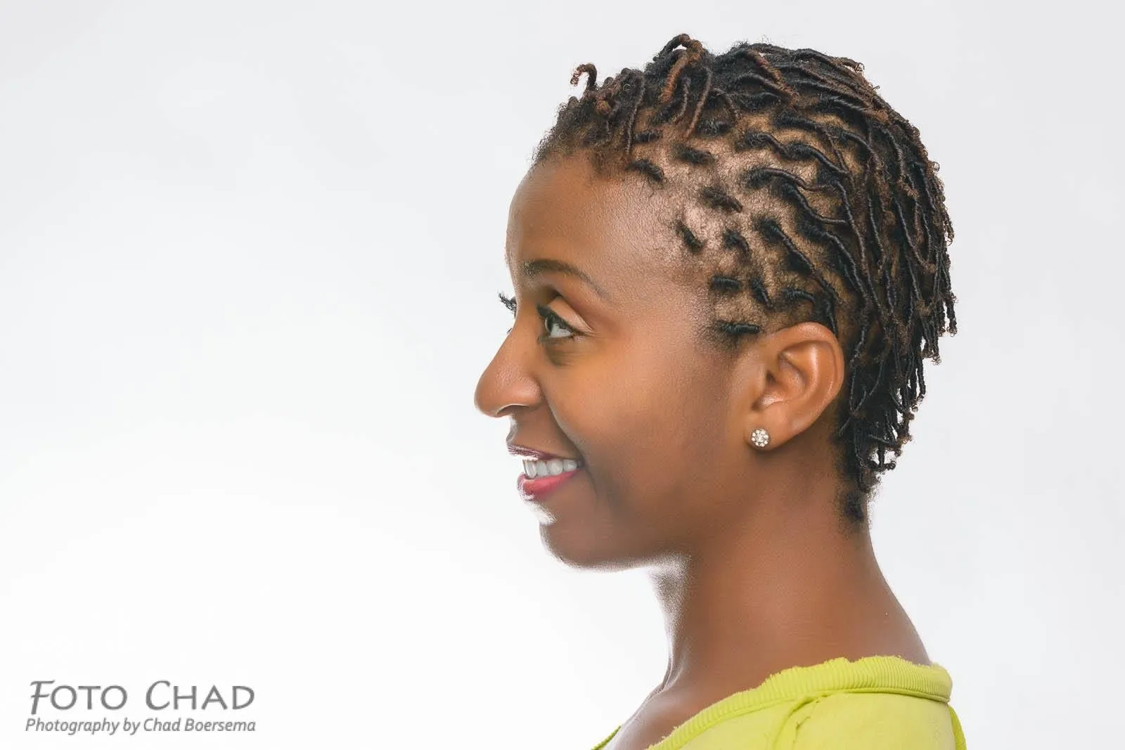 Side profile of a smiling woman with short coiled hair with caramel-tinted tips against a white backdrop, styled by Tamesha Scott
