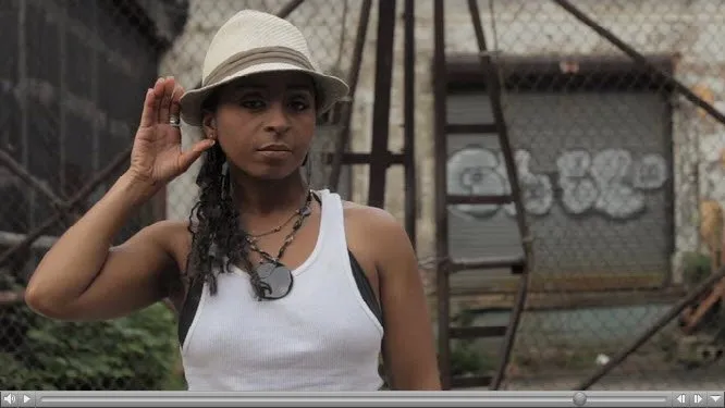 Video still of a woman in a white fedora and tank top holding a hand to her ear in front of a chain-link fence and graffiti wall.