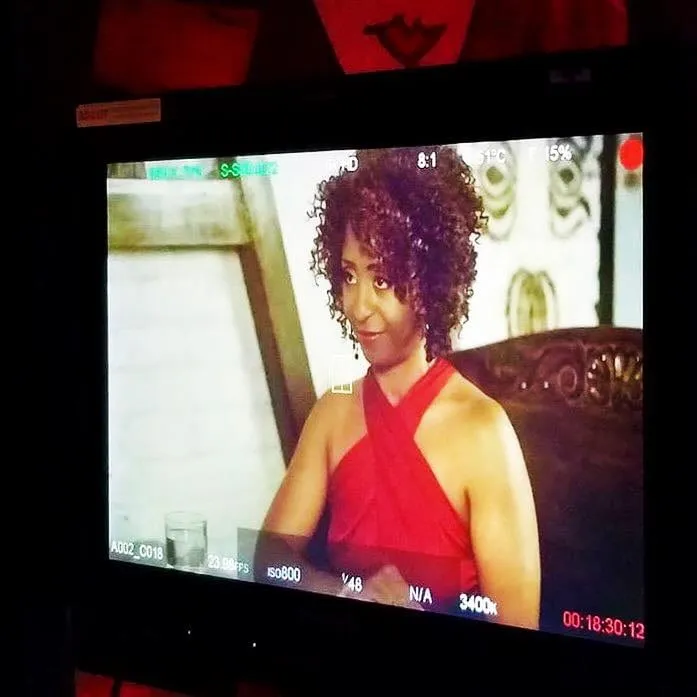 Sophia Thomas as Jasmine Jeffers in a camera-monitor screenshot from It’s a Date: a woman with short curly hair in a red halter top at a restaurant table.