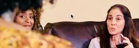 Cropped, blurry screenshot from The Mommy Group Debacle: two women on a brown leather couch, one speaking while holding a wine glass