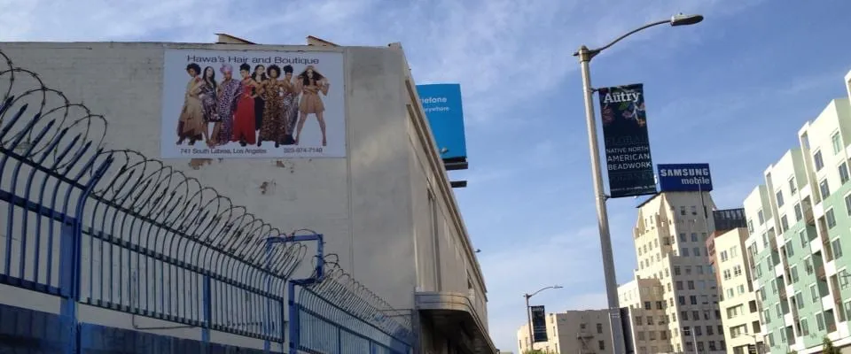 The Hawa’s Hair and Boutique billboard high on a building wall on La Brea, seen from the street past a barbed-wire fence and street banners
