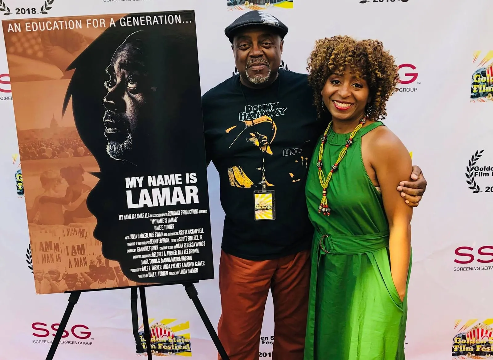 Sophia Thomas in a green dress hugs Dale E. Turner, in a flat cap, beside the My Name Is Lamar poster at the Golden State Film Festival.