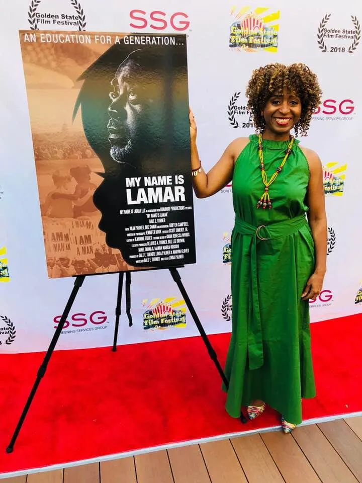 Smiling woman in a green halter dress and beaded necklace stands beside the My Name Is Lamar poster on a Golden State Film Festival red carpet.
