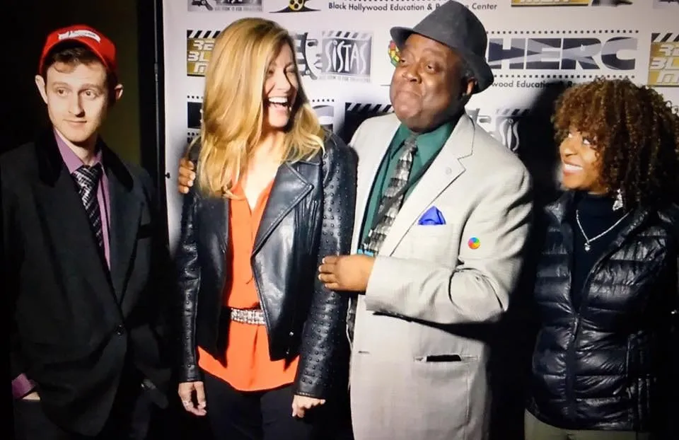 Four of the film’s cast laughing together in front of the Black Hollywood Education & Resource Center backdrop, a man in a gray blazer and hat at center.
