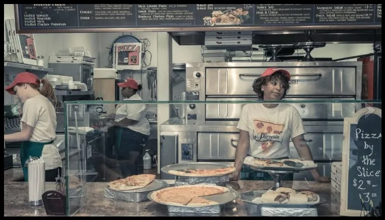 Sophia Thomas as Teresa, looking bored in a red cap and Pizzeria T-shirt behind the pizza counter in a still from My Name Is Lamar.