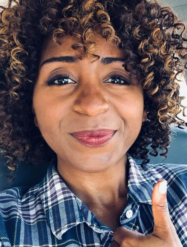 Sophia Thomas takes a smiling selfie in a blue plaid shirt, giving a thumbs up.