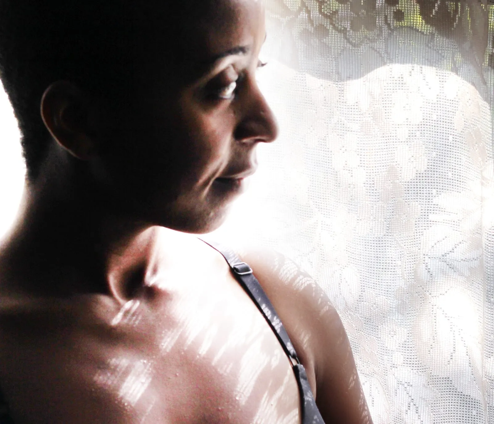 Backlit profile of a woman with cropped hair looking down beside a sunlit lace curtain, with shadow patterns on her shoulder